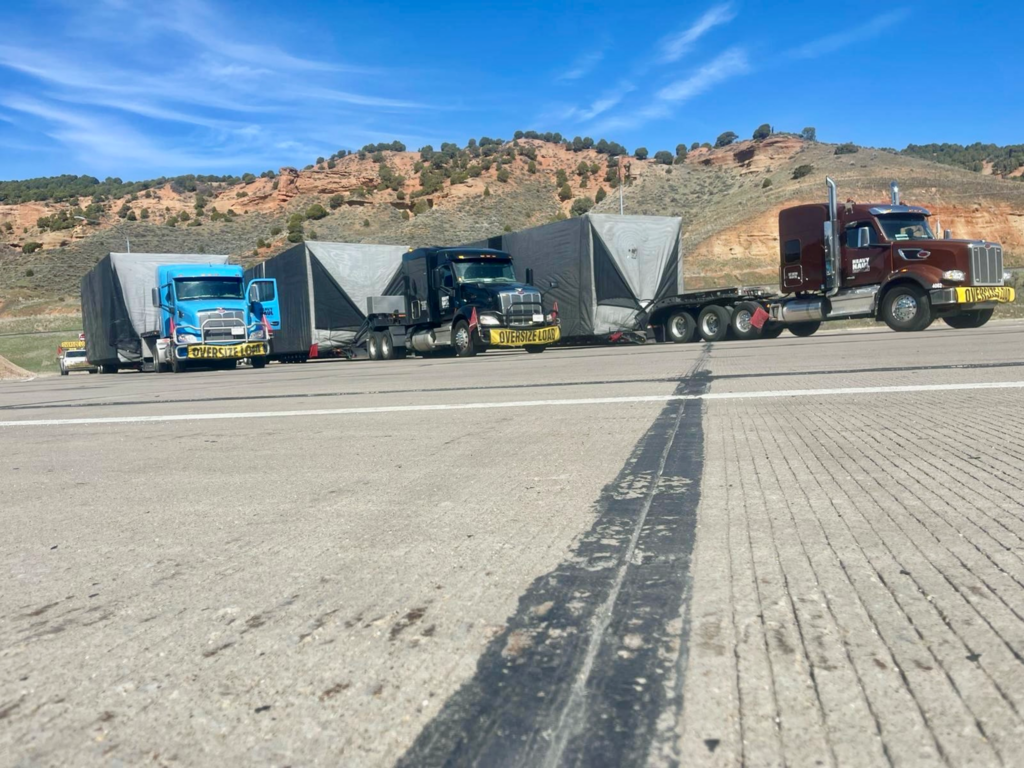 Oversize load convoy of heavy haul trucks hauling tarped superloads on flatbed trailers.