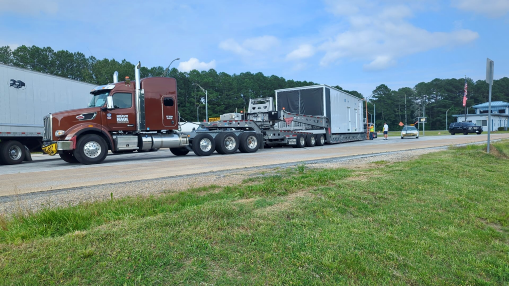 Oversize load transport hauling a prefabricated building enclosure on a heavy-duty trailer.