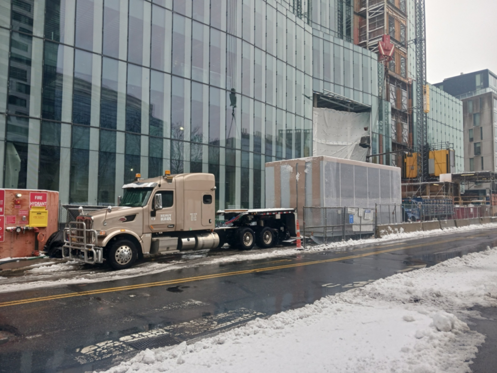 Heavy haul tractor parked curbside with a shrink-wrapped equipment skid load at an urban construction project with cranes and snow.