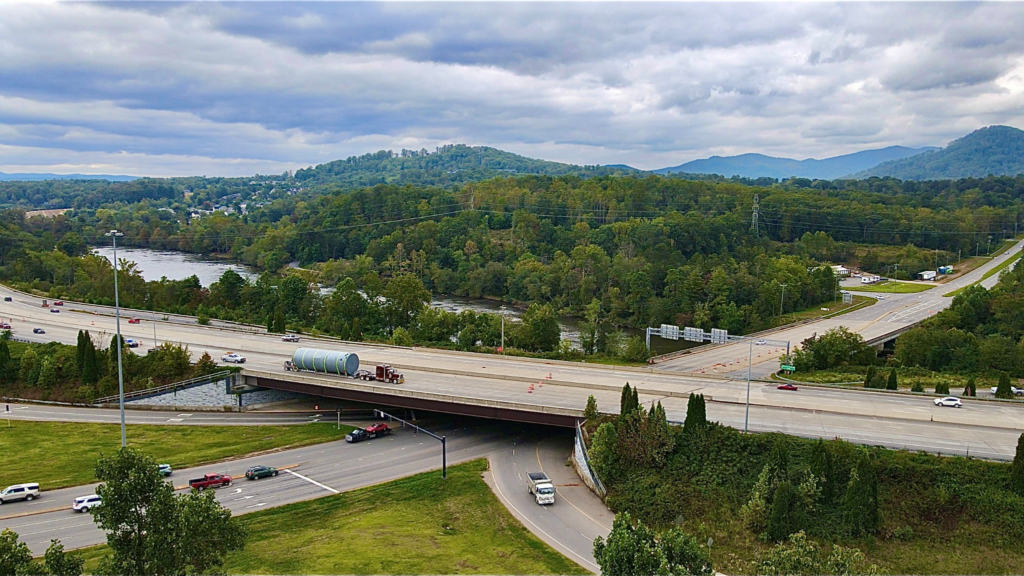 Oversize load tank hauling on a multi-axle trailer crossing a highway bridge in mountainous terrain.